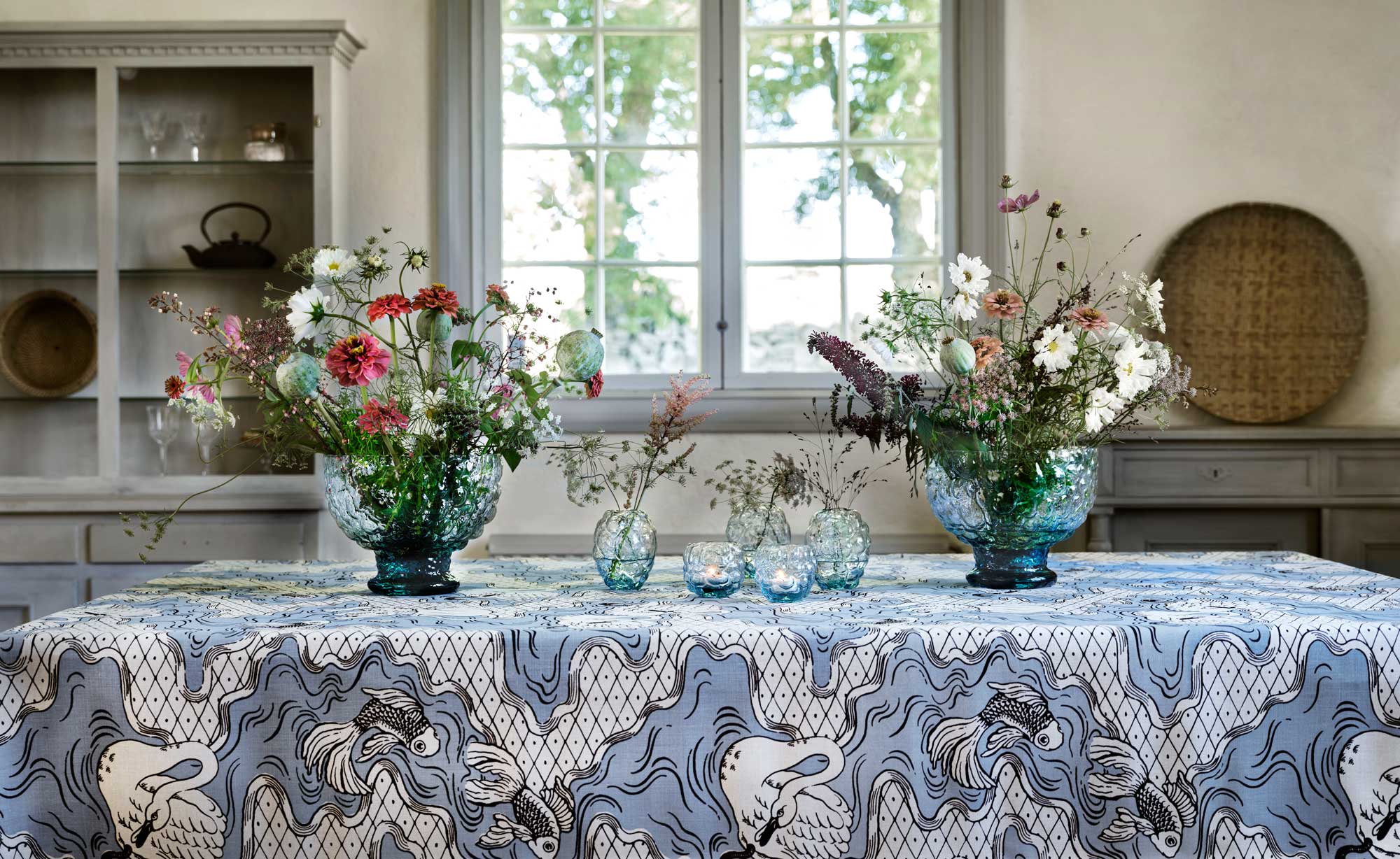 Stor bordsduk uppsydd av mönstret Opera fantasia i blå och brun färgställning. Matrum med blommor i vaser och ljusstakar på duken.